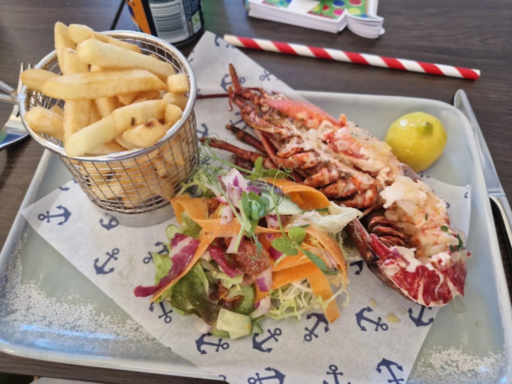 Halved grilled lobster with salad, a lemon and a basket of chips on anchor-print paper on a tray