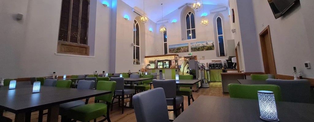 Tall restaurant hall with arched windows, blue uplighting, green and grey chairs, and a bar at the far end