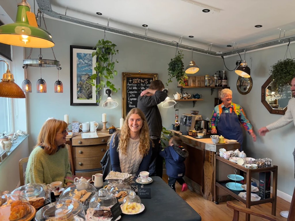 Customers smiling at a table of cakes under glass domes in a cosy café, with a woman in an apron by the counter