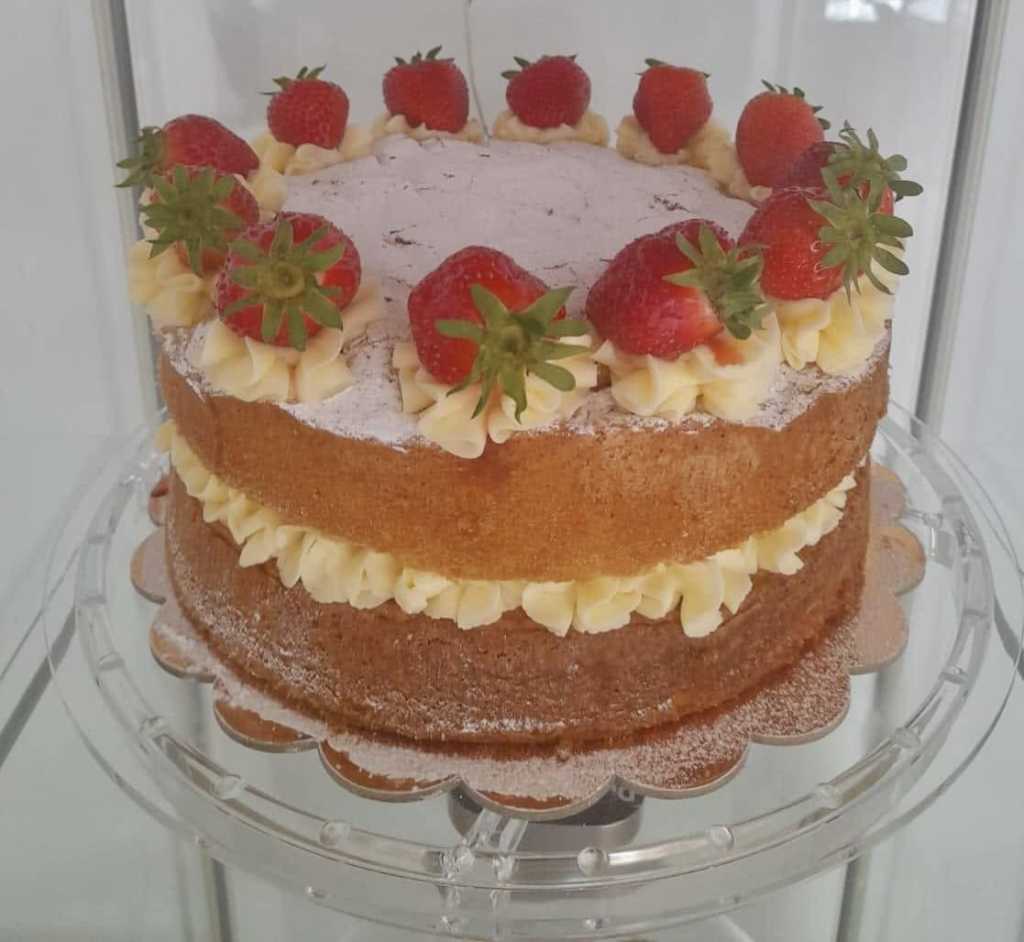Victoria sponge cake filled with buttercream, dusted with icing sugar and topped with strawberries on a glass cake stand