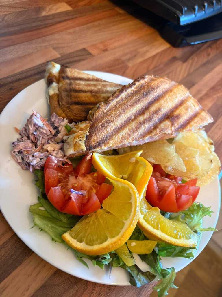 Toasted panini with coleslaw, crisps, tomato, orange slices and salad leaves on a white plate