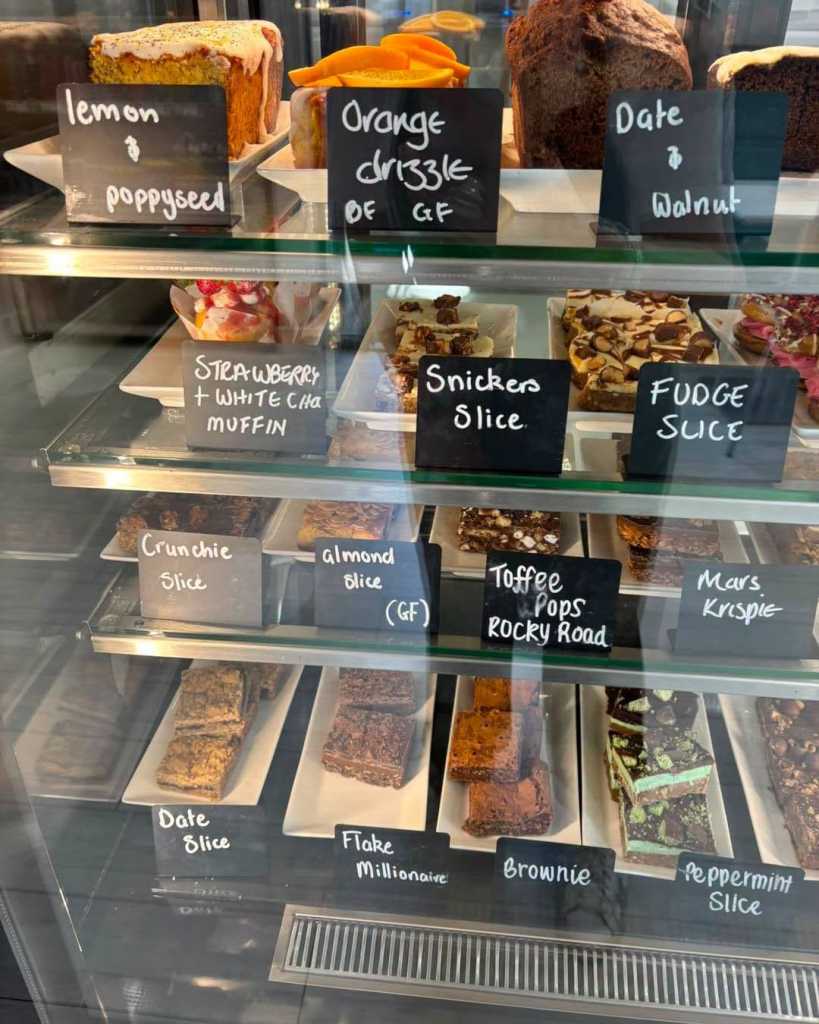 Glass cabinet of cakes and traybakes with chalk labels such as lemon and poppyseed, fudge slice and brownie