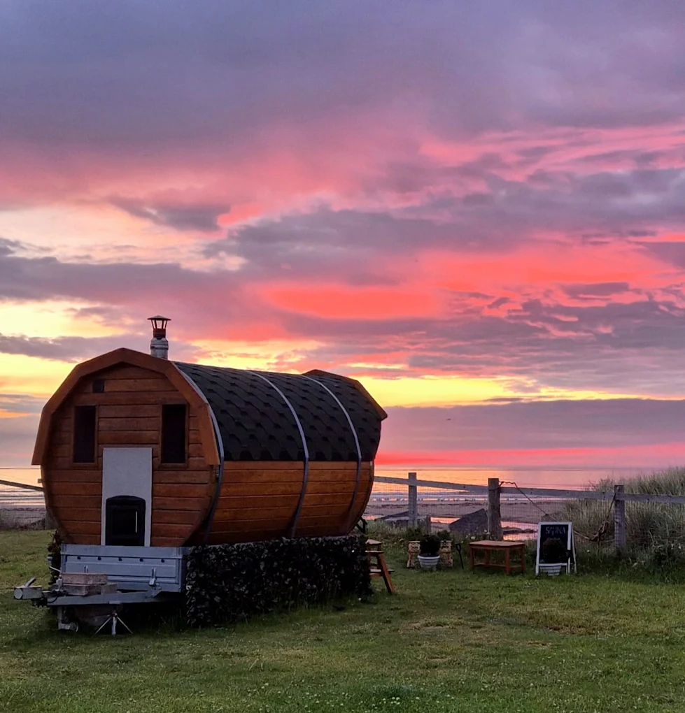 Barrel-shaped wooden sauna on a grassy clifftop by the sea under a pink and orange sunset sky