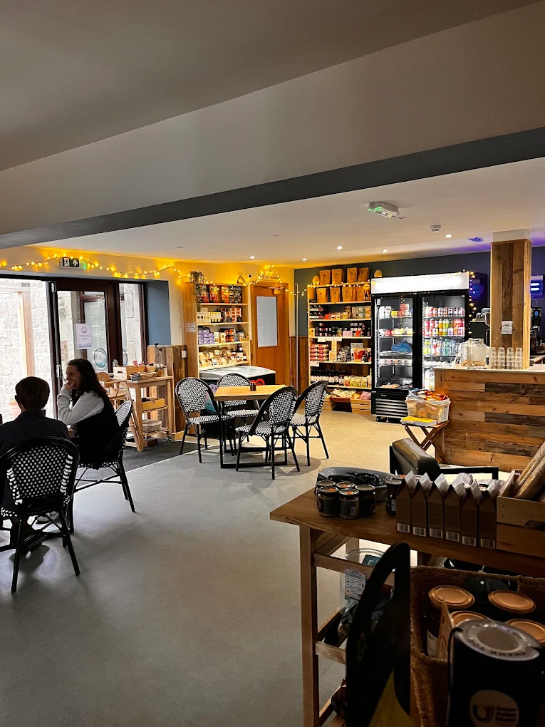 Café interior with tables and chairs, shelves of deli goods, a drinks fridge and a wooden counter under fairy lights