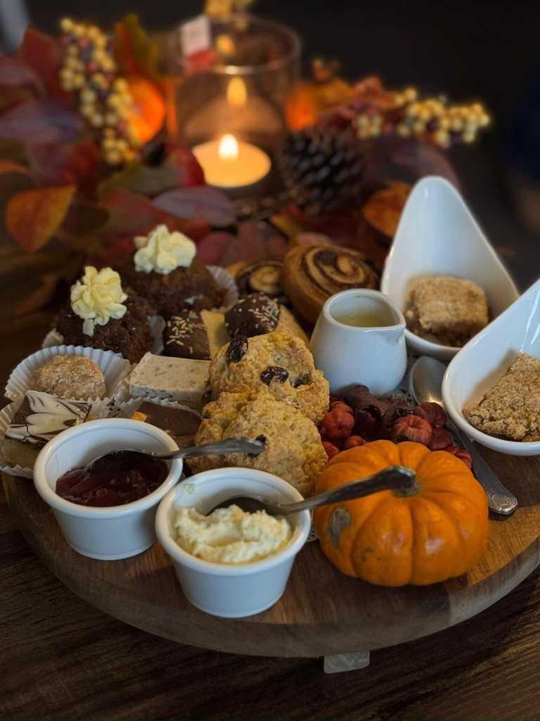Wooden board of scones, cakes, traybakes, jam and cream with a small pumpkin, autumn leaves and a candle behind