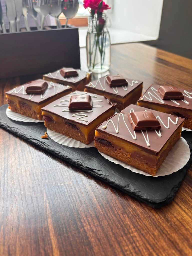 Six squares of caramel shortbread topped with chocolate, white drizzle and a chocolate piece, on a slate board