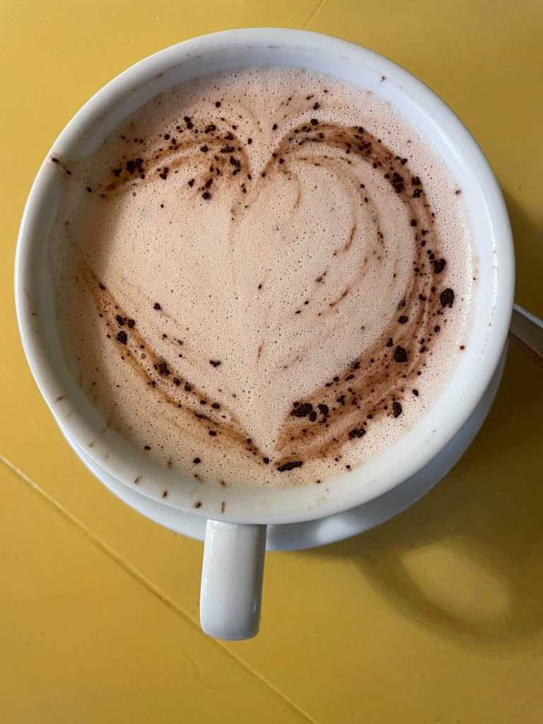 Hot chocolate in a white cup and saucer with a chocolate heart drawn in the froth, on a yellow table