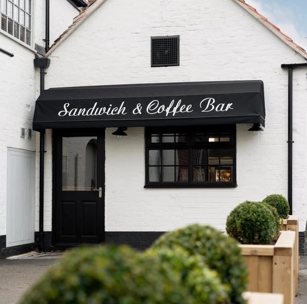 White building with a black awning reading Sandwich & Coffee Bar above a door and window, with box hedges in planters