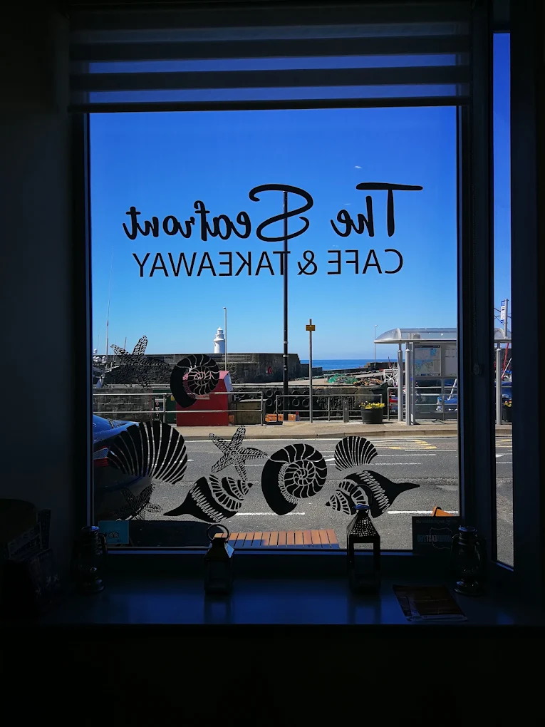 Window reading The Seafront Cafe & Takeaway with shell decals, looking out on a harbour wall and white lighthouse