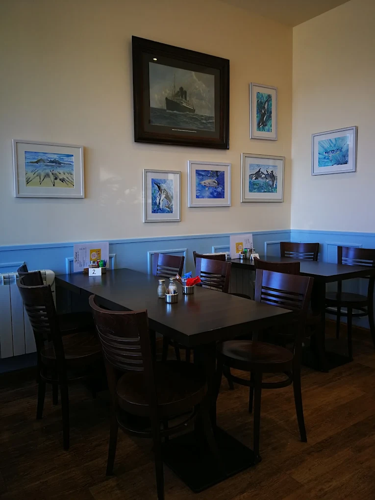 Café seating with dark wooden tables and chairs below framed sea paintings, including a steamship and marine life